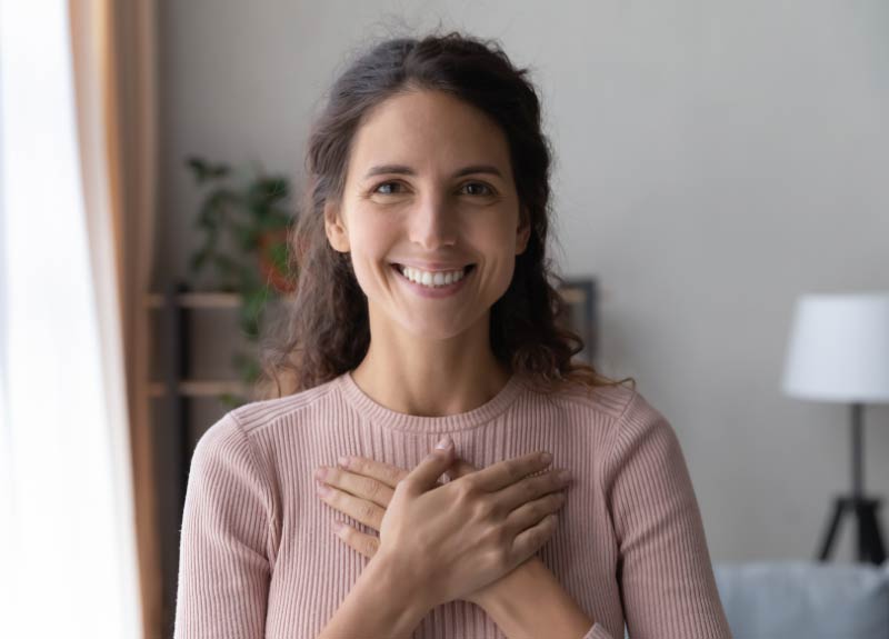 A woman in a pink sweater smiling at the camera and holding both of her hands on her heart